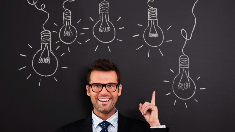 Happy man in glasses pointing at light bulbs on a chalkboard, showing creative investing ideas.